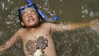 A boy lies on his back in the water, covered in shells. 