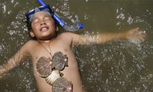 A boy lies on his back in the water, covered in shells. 